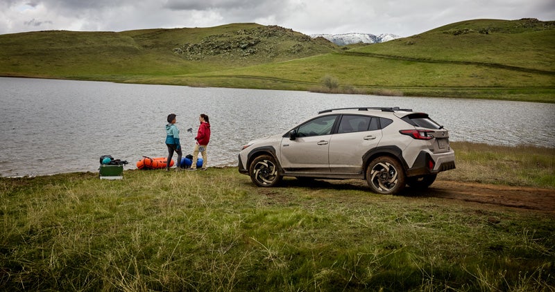A Subaru Crosstrek Hybrid parked beside a lakeshore on a grassy field, with two people standing near camping gear by the water under a cloudy sky and rolling green hills.