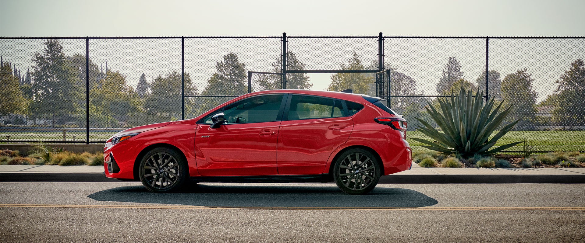 Subaru Impreza car parked on a city street with a chain-link fence and greenery in the background.