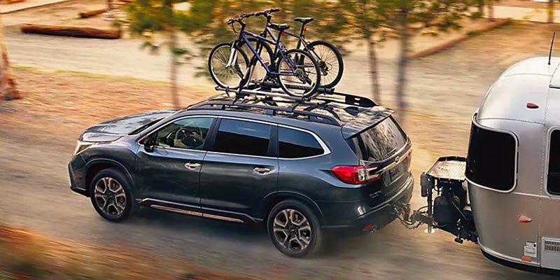 A dark gray SUV carrying bicycles on a roof rack and towing a silver travel trailer along a dirt road.
